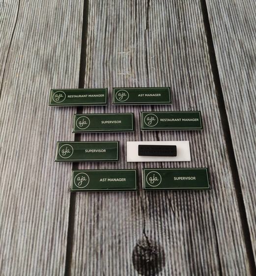 Bottle green name badges with white logo and staff role titles, eight laid on grey wood, custom made for AJA by RestroCart, New Delhi