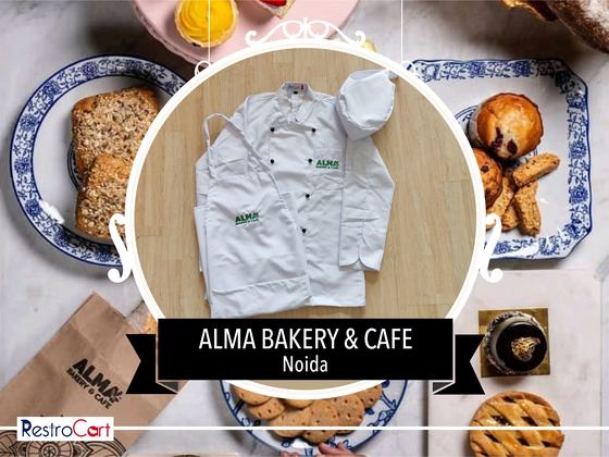 White chef coat with black buttons and green logo, white chef cap and bib apron, laid flat, custom made for Alma Bakery by RestroCart, New Delhi