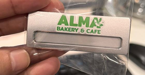 Silver metal name badge with green printed logo and slot window, held in hand, custom made for Alma Bakery by RestroCart, New Delhi