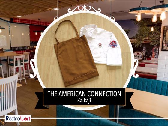 Brown canvas bib apron beside a folded white shirt with round embroidered logo, custom made for American Connection by RestroCart, New Delhi