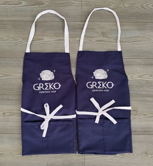 Two navy bib aprons with white neck straps and ties and white embroidered logo, custom made for Greko Espresso Bar by RestroCart, New Delhi