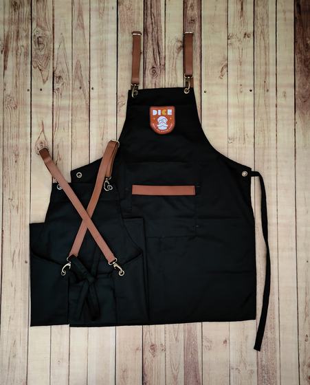 Black canvas bib apron with tan leather cross-back straps, leather pocket trim and orange badge logo, custom made by RestroCart, New Delhi