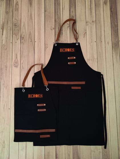 Black canvas apron pair with tan leather straps, pocket trims and orange logo patch, custom made by RestroCart, New Delhi