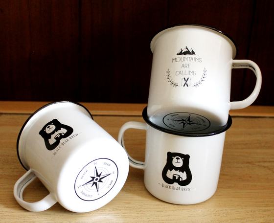 White enamel mugs with black rims, bear mascot and compass print, three stacked, close view, custom made for Black Bear Brew by RestroCart, New Delhi
