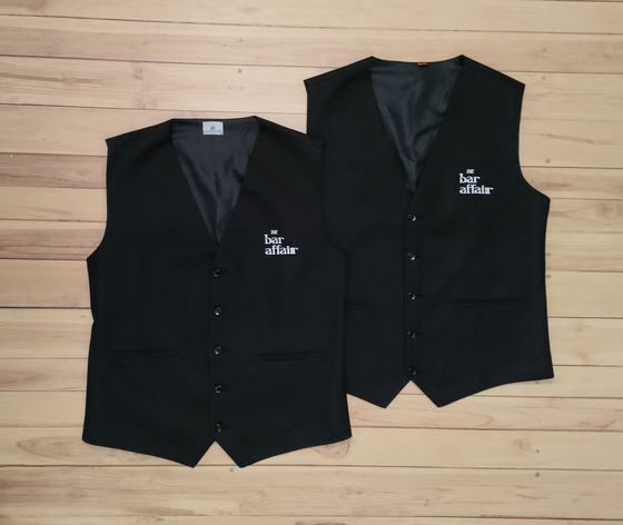 Black waistcoats with white embroidered logo on chest, front view pair on wood, custom made by RestroCart, New Delhi
