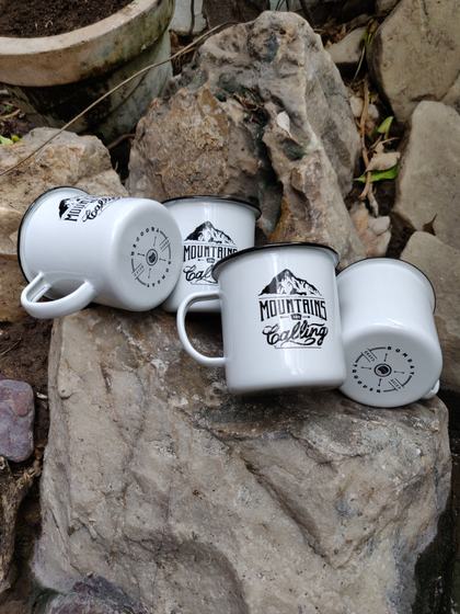 White enamel mugs with black mountain print and slogan, four resting on rocks outdoors, custom made by RestroCart, New Delhi