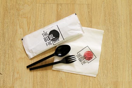 Paper cutlery pouch and printed napkin with red logo beside black plastic spoon and fork, custom made by RestroCart, New Delhi