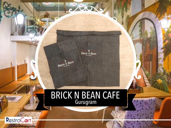 Two charcoal denim waist aprons with small printed logo, laid flat on a wooden table, custom made for Brick N Bean Cafe by RestroCart, New Delhi