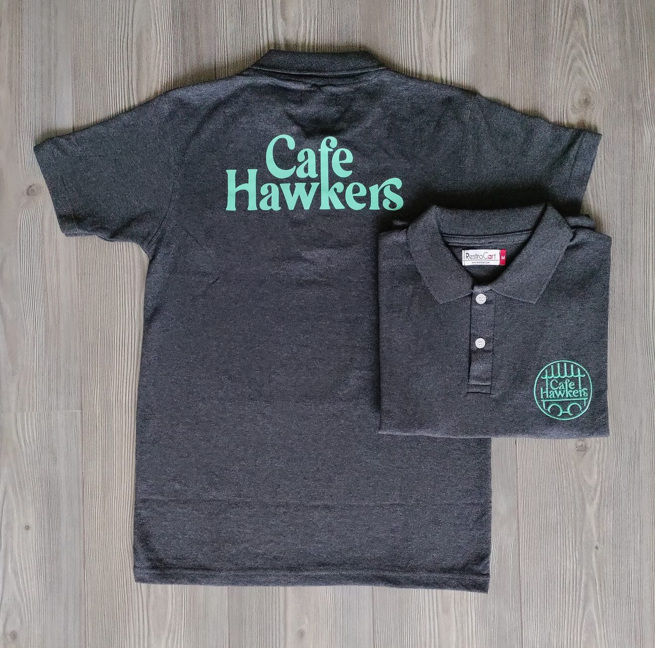 Charcoal polo t-shirt uniforms with a large mint print across the back and a round badge logo on the chest, made for Cafe Hawkers by RestroCart