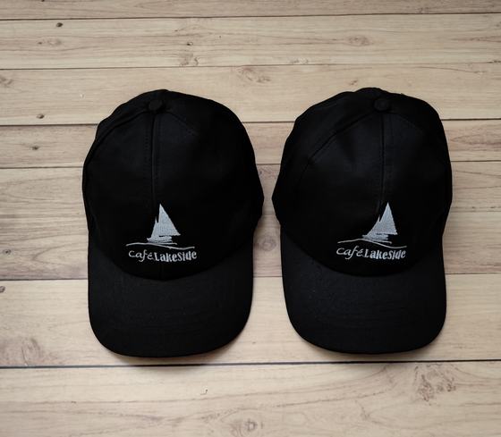 Black baseball caps with white embroidered sailboat logo, pair on light wood, custom made for Cafe Lakeside by RestroCart, New Delhi