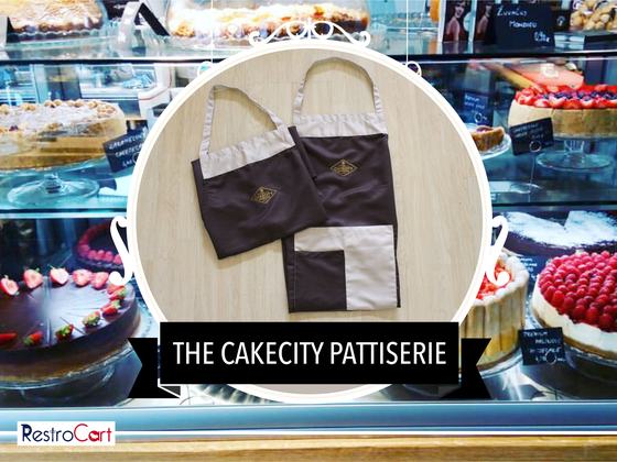 Dark brown bib aprons with beige panels and small gold logo, one folded, laid flat, custom made for CakeCity Patisserie by RestroCart, New Delhi