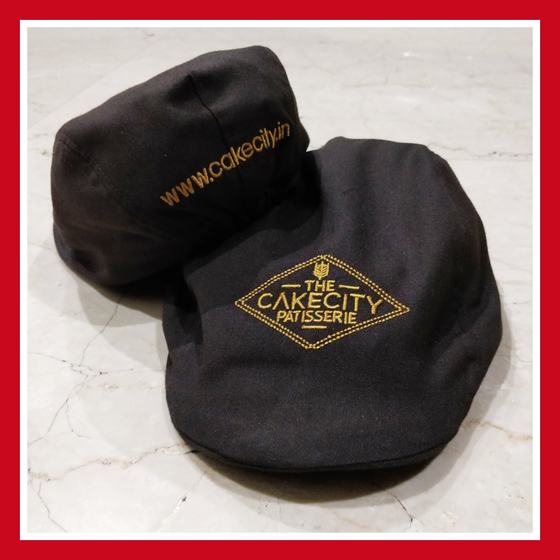 Two dark brown flat caps with gold embroidered diamond logo and web address, close-up, custom made for CakeCity Patisserie by RestroCart, New Delhi