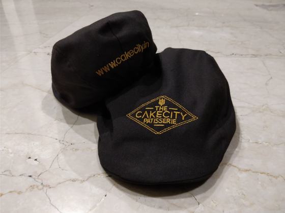 Dark brown flat caps with gold embroidered diamond logo and website text, on marble, custom made for CakeCity Patisserie by RestroCart, New Delhi