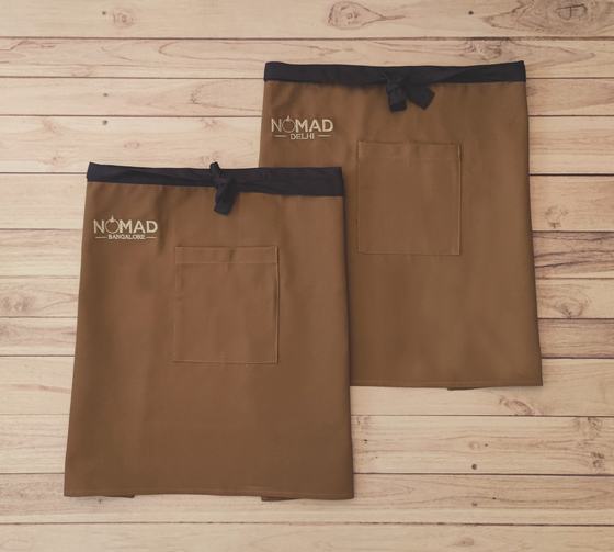 Two brown canvas waist aprons with black waistband and ties and white printed logo on the pocket, custom made by RestroCart, New Delhi