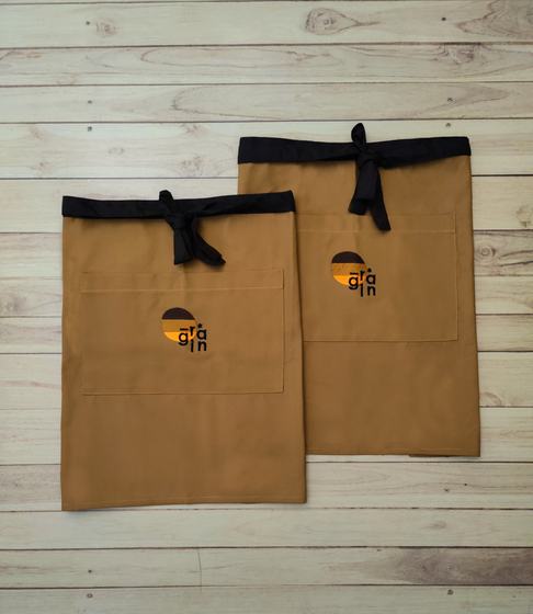 Two mustard canvas waist aprons with black waistband and ties and orange and black round logo, custom made by RestroCart, New Delhi