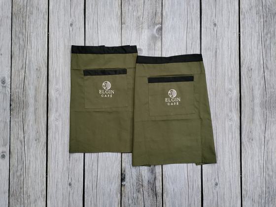 Two olive canvas waist aprons with black waistband and pocket trim and white printed logo, on grey wood, custom made by RestroCart, New Delhi