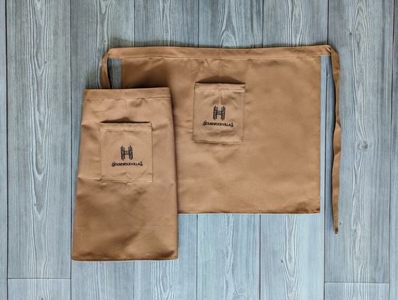 Tan canvas long waist apron and short waist apron with pockets and black printed logo, on grey wood, custom made by RestroCart, New Delhi