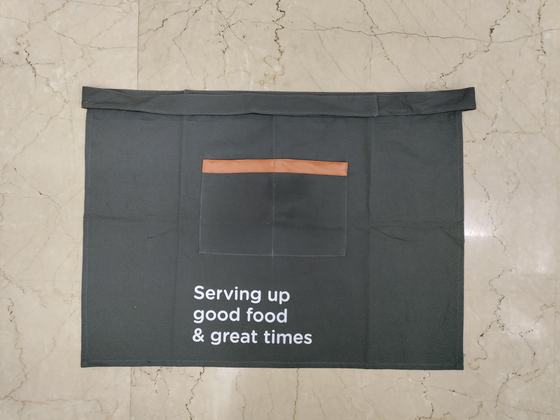 Black canvas waist apron with tan leather trimmed pocket and white printed slogan, laid on marble, custom made by RestroCart, New Delhi