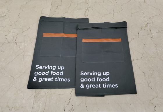 Two black canvas waist aprons with tan leather pocket trims and white printed slogan, overlapping, custom made by RestroCart, New Delhi