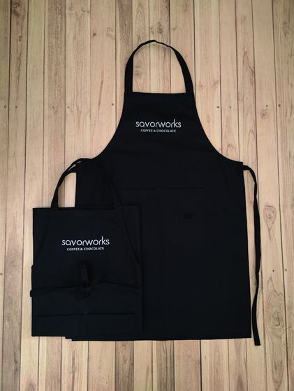 Two black canvas bib aprons in large and small sizes with white printed chest logo, laid flat, custom made by RestroCart, New Delhi