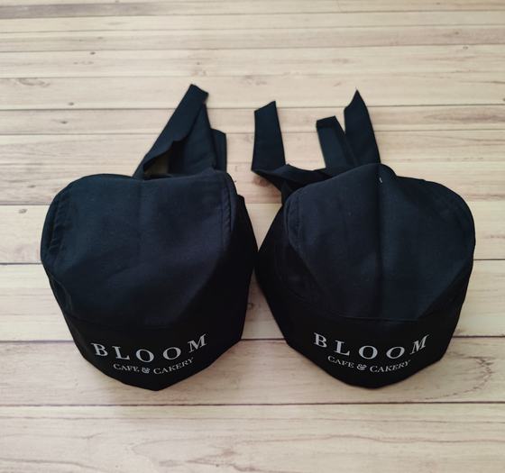 Pair of black chef skull caps with long back ties and white printed cafe and cakery wordmark, custom made by RestroCart, New Delhi