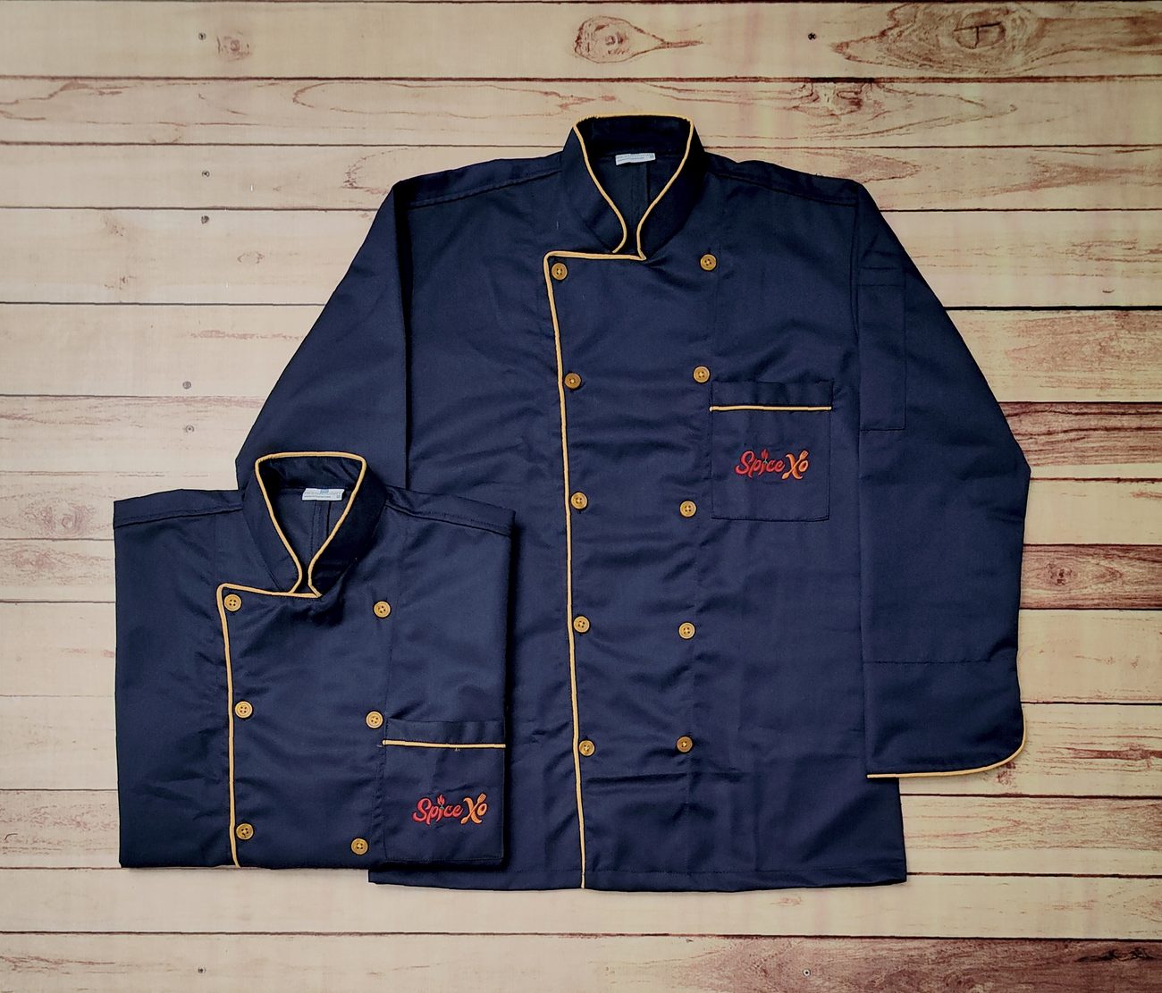 Navy double-breasted chef coats with gold contrast piping, two rows of gold buttons and embroidered branding on the chest, laid flat on wooden boards