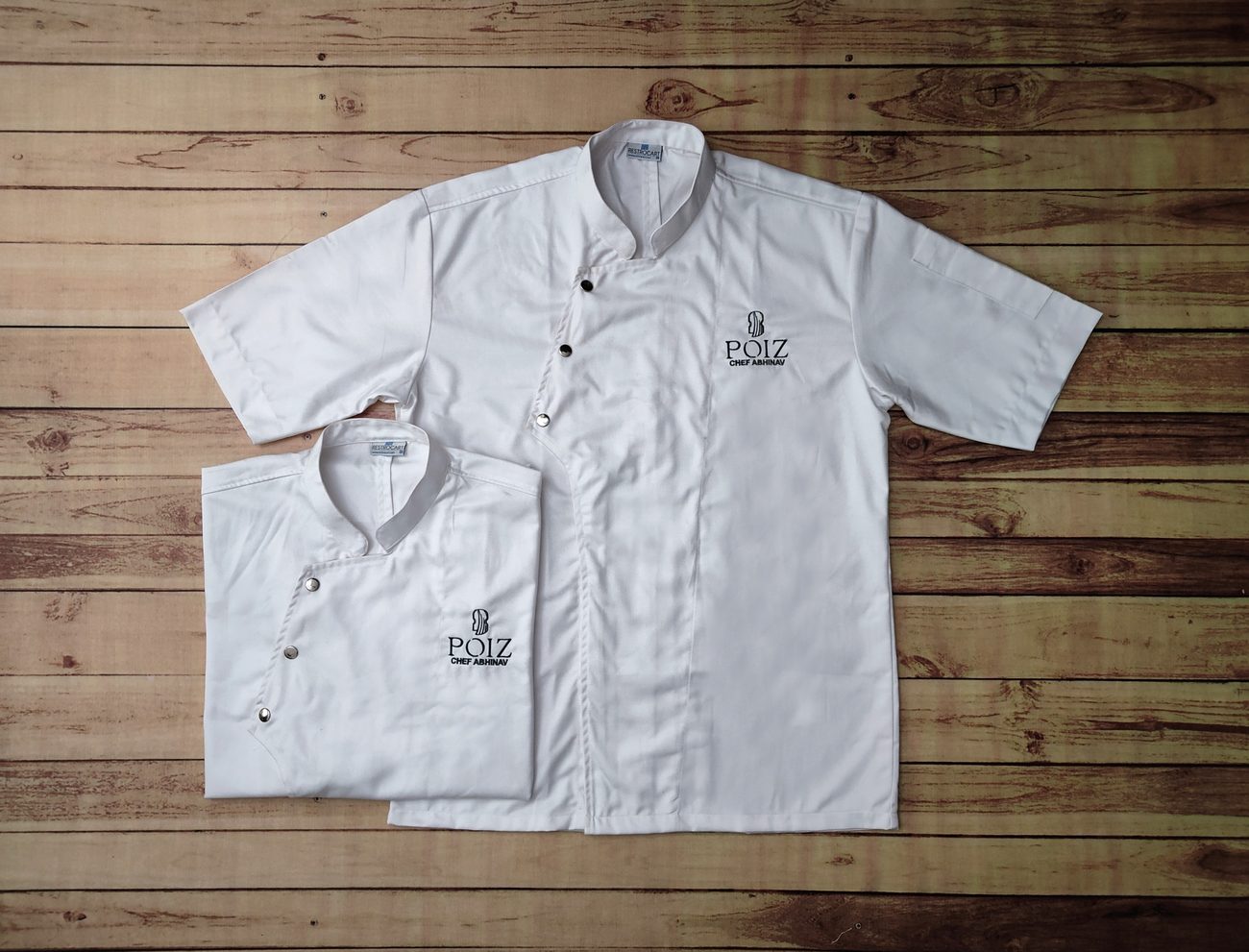 White half-sleeve chef coats with an offset front, black press studs and small embroidered branding on the chest, laid flat on wooden boards