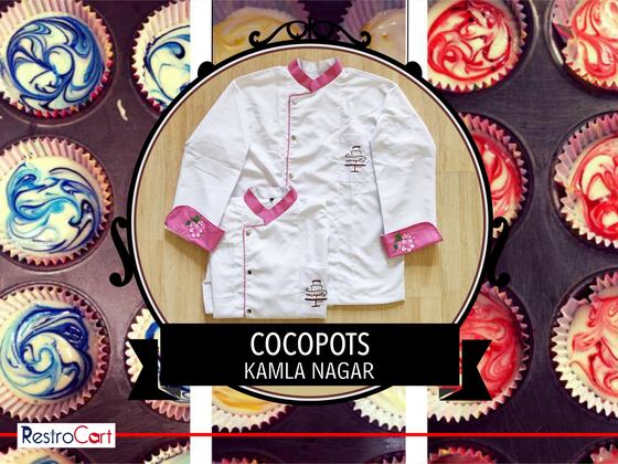 Two white chef coats with pink collar, pink cuffs and printed cake logo, laid flat, custom made for Cocopots by RestroCart, New Delhi