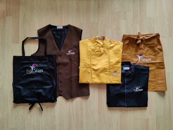 Set of folded black bib apron, brown waistcoat, mustard chef coat, navy shirt and tan waist apron with logos, custom made by RestroCart, New Delhi