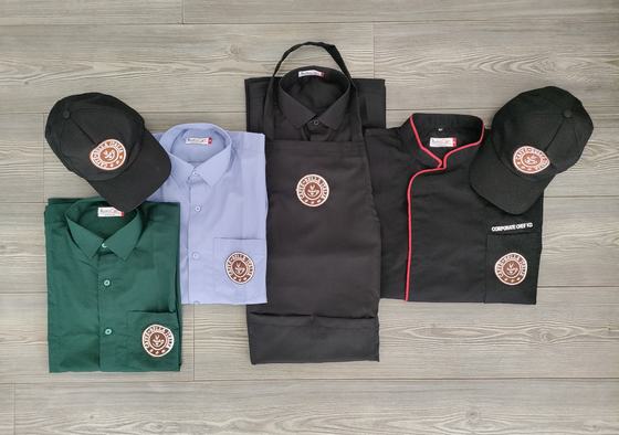 A complete branded uniform set laid out together: green and blue shirts, black apron, black chef coat with red piping and black caps, all carrying the same round logo