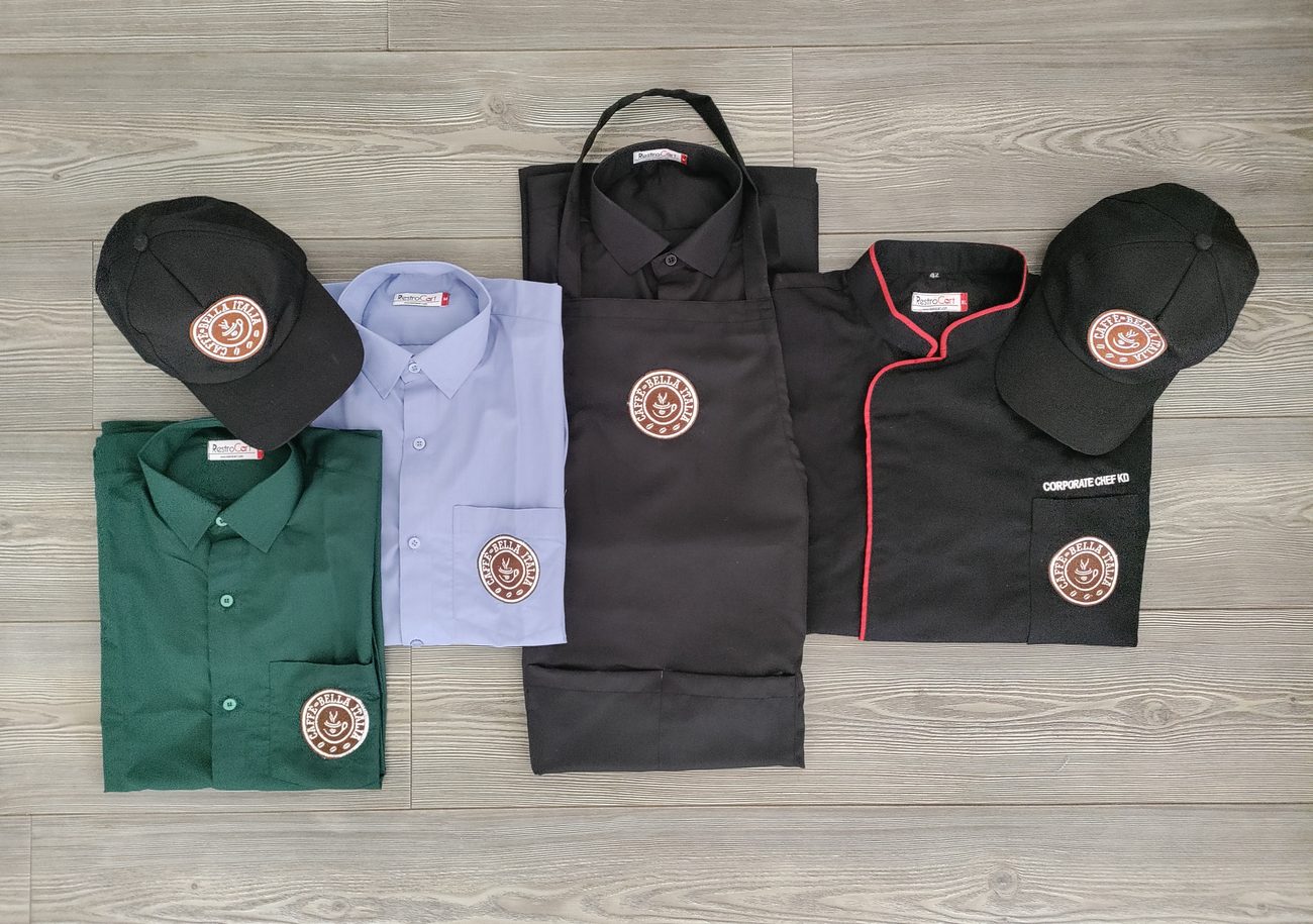 A complete branded uniform set laid out together: green and blue shirts, black apron, black chef coat with red piping and black caps, all carrying the same round logo