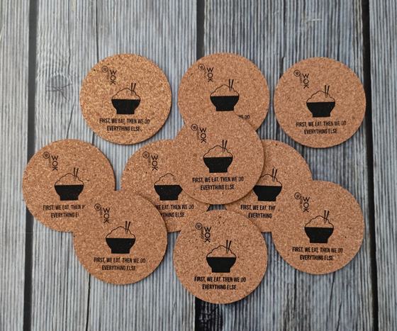 Set of round cork coasters with black printed noodle bowl logo and slogan, scattered on grey wood, custom made by RestroCart, New Delhi