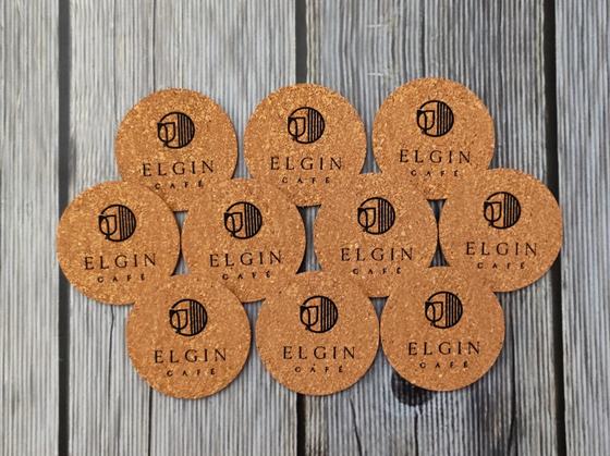 Set of ten round cork coasters with black printed cafe logo and wordmark, arranged on grey wood, custom made by RestroCart, New Delhi