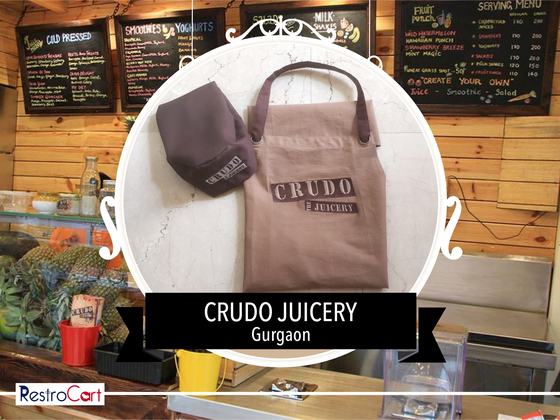 Brown canvas bib apron with printed logo and matching brown cap, on a white counter, custom made for Crudo Juicery by RestroCart, New Delhi