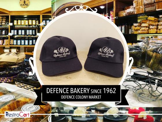 Two navy baseball caps with white embroidered monogram logo, on a wooden table, custom made for Defence Bakery by RestroCart, New Delhi