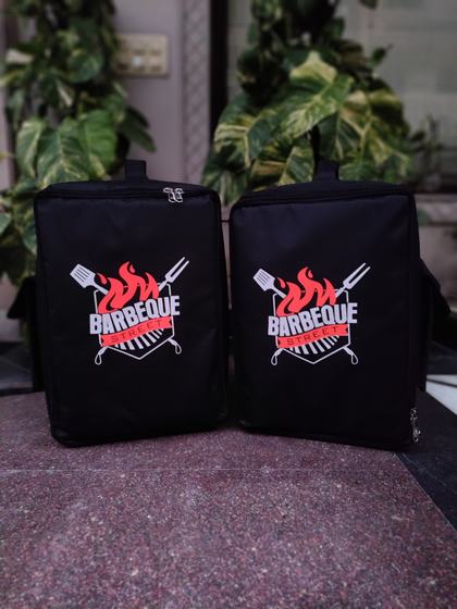 Two black insulated food delivery backpacks with red and white printed grill logo, standing closed, custom made by RestroCart, New Delhi