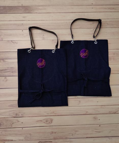 Two dark denim bib aprons with leatherette straps and purple and pink embroidered round logo, custom made by RestroCart, New Delhi
