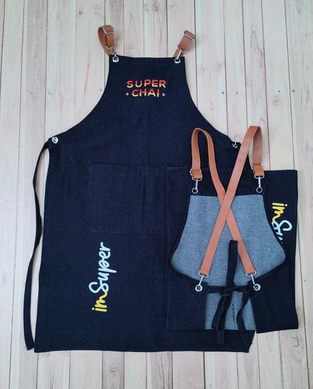 Large denim bib apron with colourful printed text beside grey denim apron with crossed tan leatherette straps, custom made by RestroCart, New Delhi