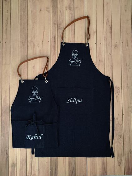 Two black denim bib aprons with tan leatherette neck straps, white logo and embroidered staff names, custom made by RestroCart, New Delhi