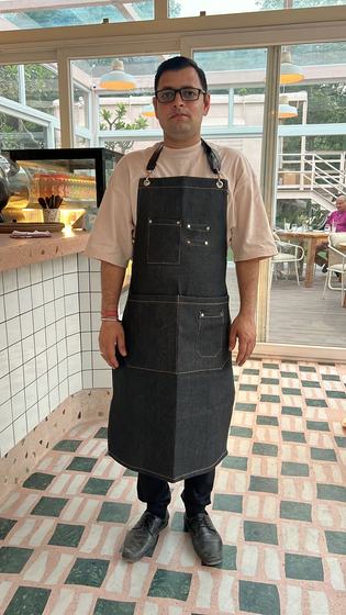 Man wearing a dark denim bib apron with tan leather straps and multiple pockets in a cafe, custom made by RestroCart, New Delhi