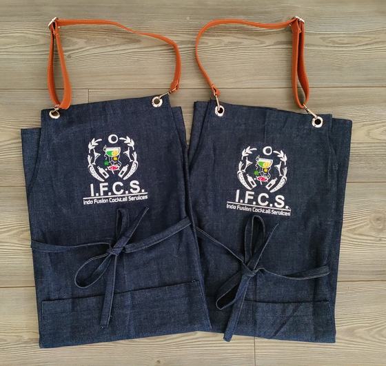 Two denim bib aprons with tan leather neck straps and white embroidered crest logo, laid side by side, custom made by RestroCart, New Delhi