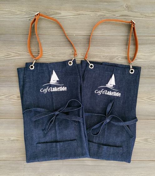 Two denim bib aprons with tan leather straps, white sailboat logo and tie waist, laid side by side, custom made by RestroCart, New Delhi