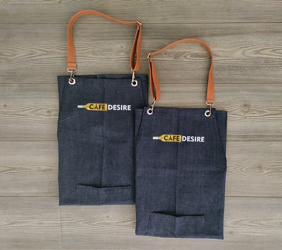 Two denim bib aprons with tan leather neck straps and yellow and white printed logo, one overlapping, custom made by RestroCart, New Delhi