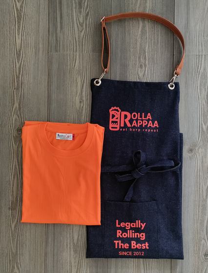 Dark denim bib apron with tan leather strap and red printed logo and slogan beside a folded orange t-shirt, custom made by RestroCart, New Delhi