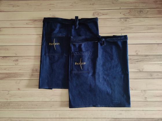 Two denim waist aprons with gold embroidered script logo and front pocket, overlapping on wooden floor, custom made by RestroCart, New Delhi