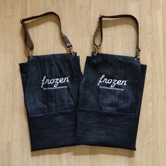 Two denim bib aprons with brown leather straps, white printed script logo and contrast pocket panel, custom made by RestroCart, New Delhi
