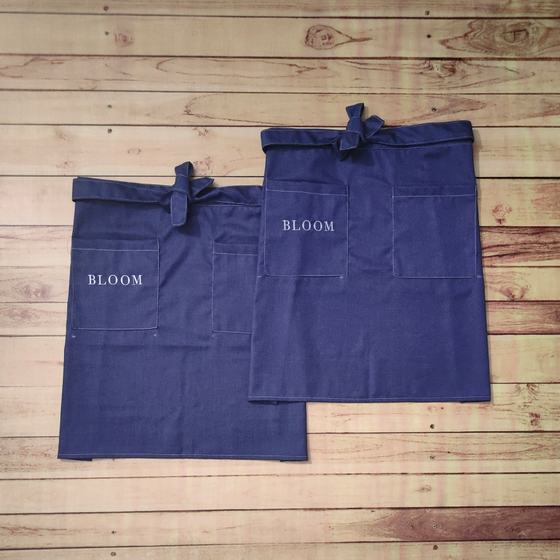 Two indigo denim waist aprons with contrast stitched pockets and small printed wordmark, laid flat, custom made by RestroCart, New Delhi