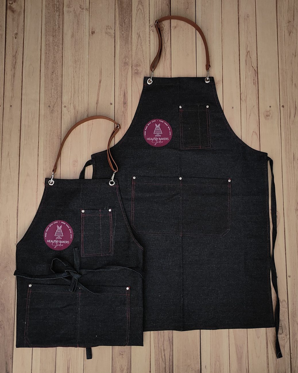 Black denim barista bib aprons with tan leather neck straps, contrast stitching and a round logo patch, custom aprons by RestroCart