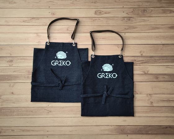Two dark denim bib aprons with black cross straps and white printed logo, laid flat, custom made by RestroCart, New Delhi