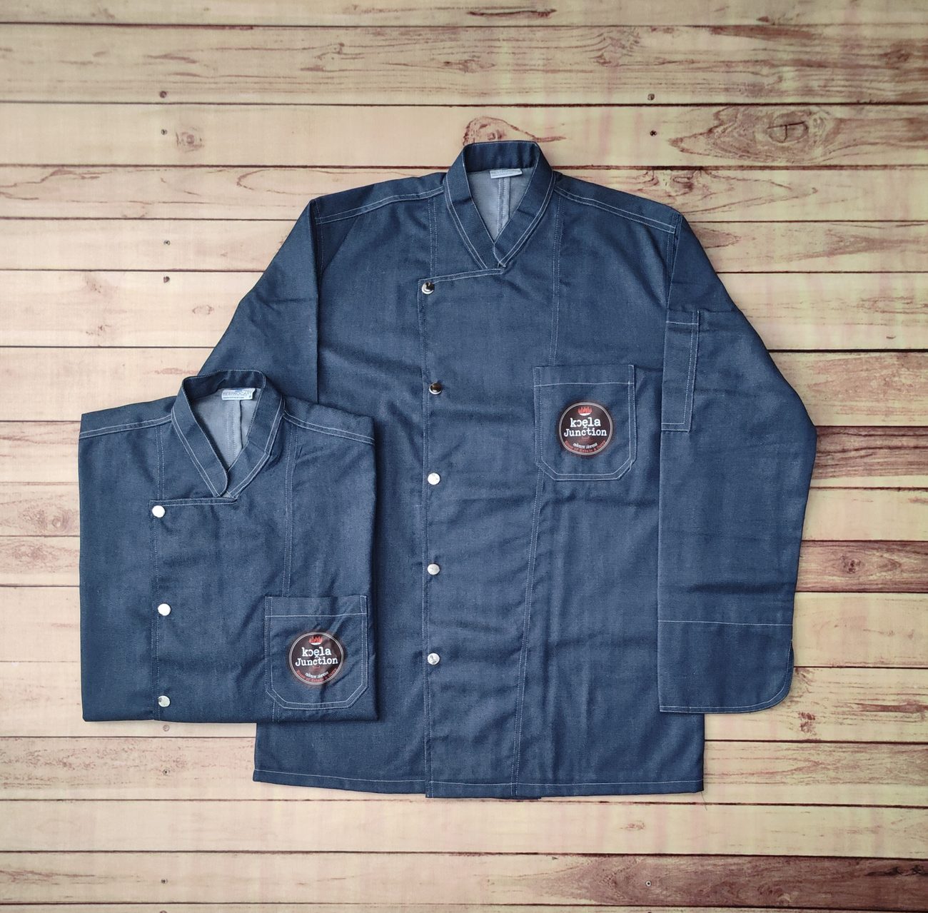Two mid-blue denim chef coats with stand collars, press studs, contrast topstitching and an embroidered round patch on the chest pocket, laid flat on wooden boards